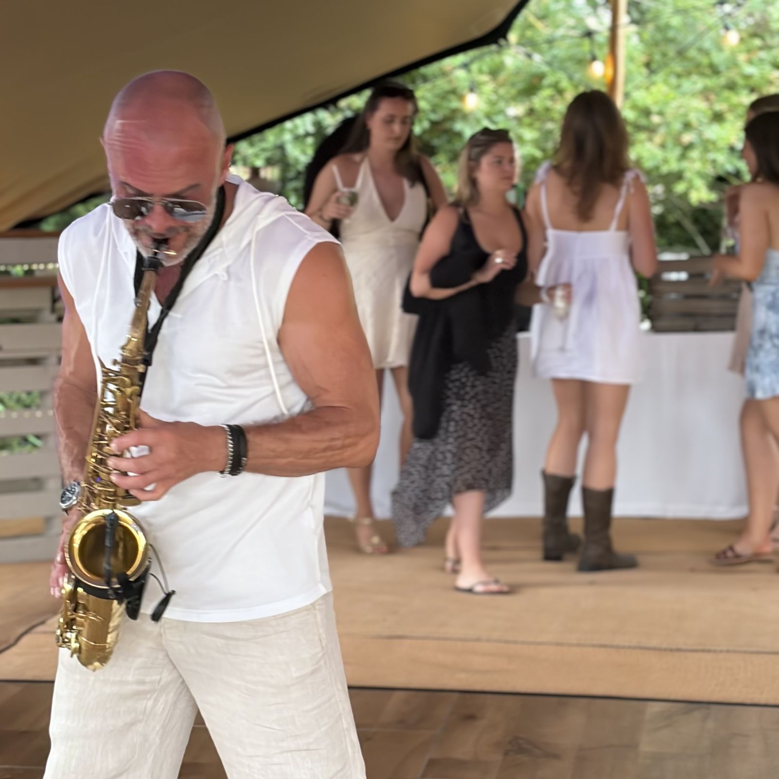 Paul Martyn playing saxophone under a marquee while guests stand talking with drinks